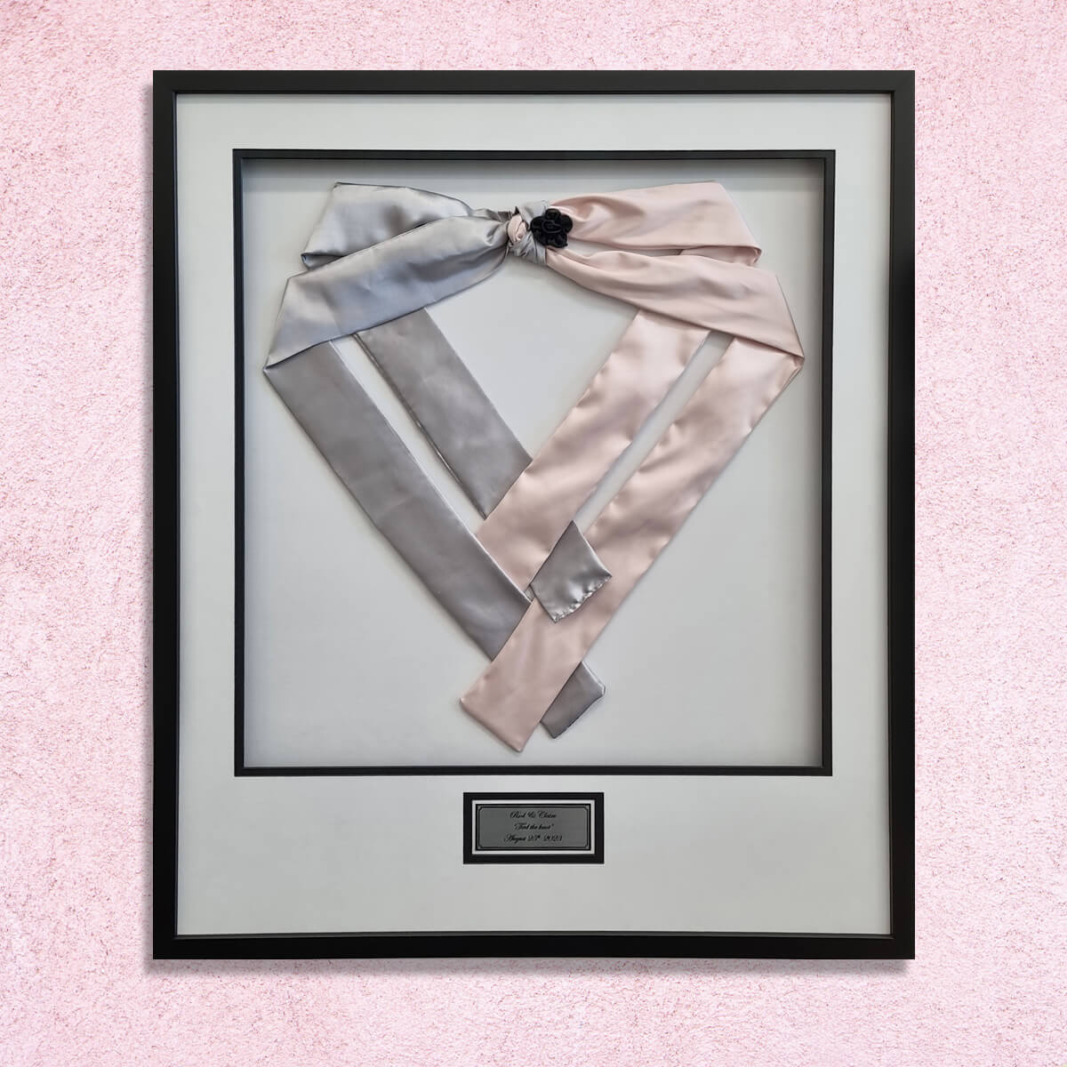 Grey and pink satin wedding sashes tied with a black rosette, pinned in a heart shape in a black frame with white double mat and engraved plaque